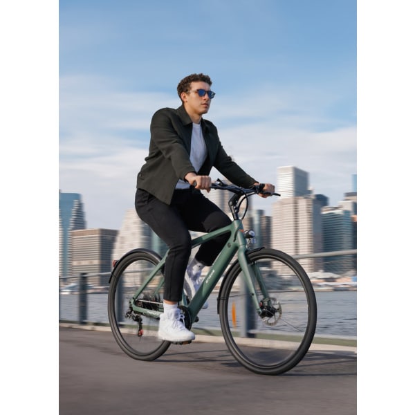 Na obrázku je muž jazdiaci na bicykli, zachytený z bočného pohľadu. Bicykel má moderný dizajn s matným povrchom v zelenej farbe. Viditeľné sú kotúčové brzdy na prednom kolese, ktoré majú strieborný lesk. Kolesá sú tenké, vhodné na mestské prostredie, s hladkým dezénom. Rám bicykla je pravdepodobne z ľahkého kovu, bez viditeľného odpruženia. Muž má na sebe tmavé oblečenie a slnečné okuliare, jazdí po ceste v mestskom prostredí s výhľadom na mrakodrapy. Fotografia je zhotovená počas dňa, čo naznačuje jasná obloha a dobré osvetlenie.
